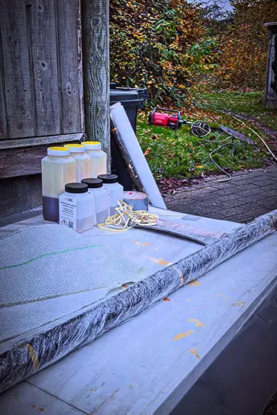 Materialer til punktrenovering af kloak ligger klar på en arbejdsflade, herunder kemikaliebeholdere, glasfiberdug og reb, med udstyr og haveområde i baggrunden